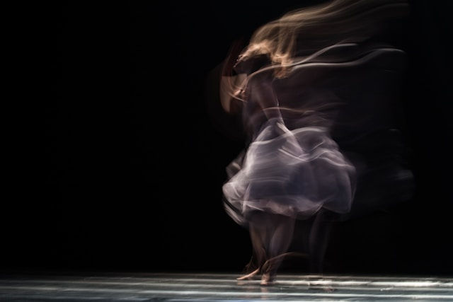 The photo captures a blurred motion of a dancer in a flowing dress, creating a dreamy and ethereal effect against a dark background.