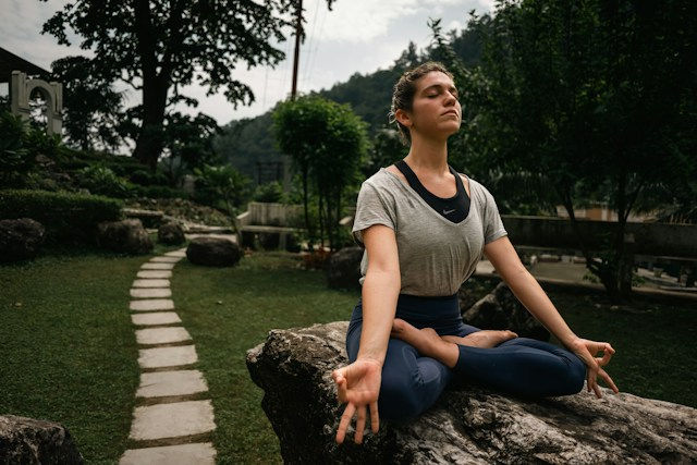 Woman meditating cross-legged on a rock in a lush garden, eyes closed, with a stone path and trees behind her.