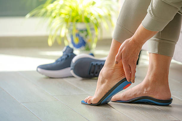 Person adjusting a blue insole on barefoot in a sunlit room with sneakers nearby, wooden floor, and a plant in a decorative pot.