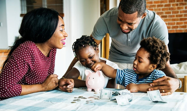 Happy family of four putting cash into a pink piggy bank at a table indoors.