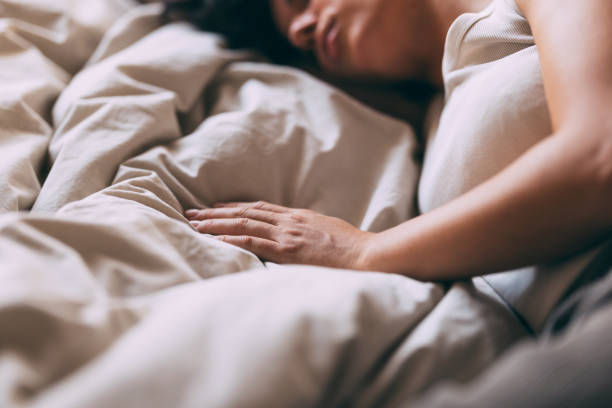 Person sleeping in a bed with beige sheets, hand resting on the blanket. Soft light creates a calm and relaxed atmosphere.