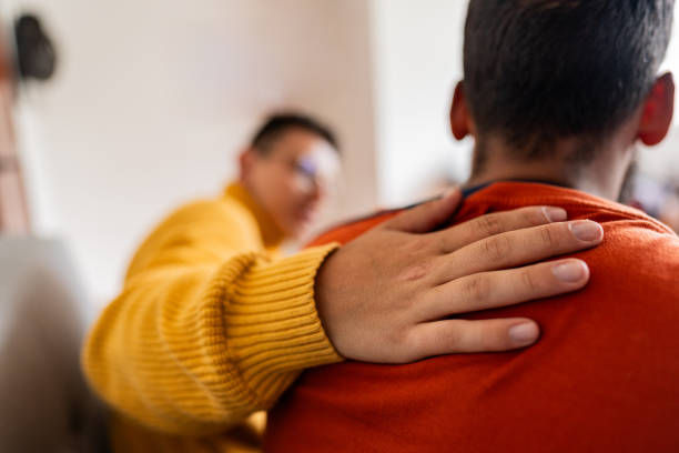 A person in a yellow sweater consoles another in an orange sweater with a comforting hand on their shoulder, indoors.