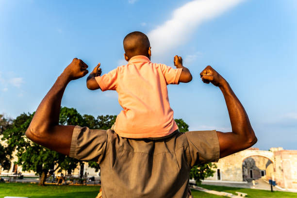 Man carrying child on shoulders in park, both flexing arms. Child in pink shirt, man in brown. Green trees and clear blue sky background.
