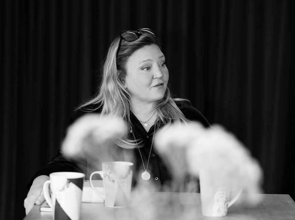 Woman at a table with coffee mugs, looking right in a black-and-white meeting scene with blurred foreground.