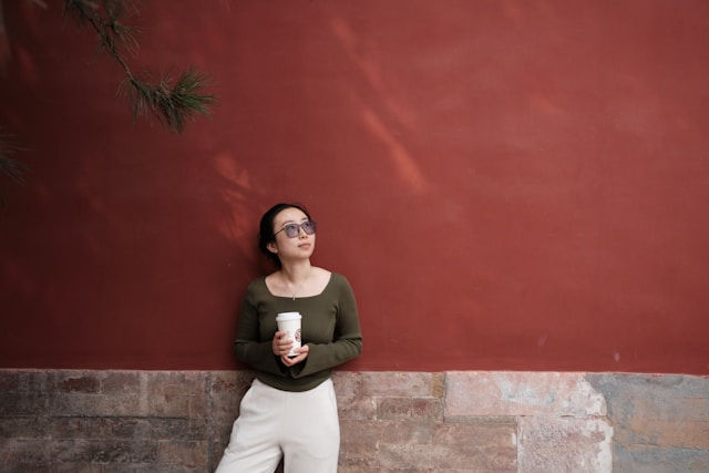 Woman in sunglasses leans against a red wall, holding a coffee cup, wearing a green top and white pants in a calm pose.