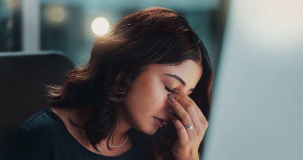 Woman in a dark shirt sits indoors, rubbing her eyes with fingers, appearing tired. Soft light in the background, creating a calm ambiance.
