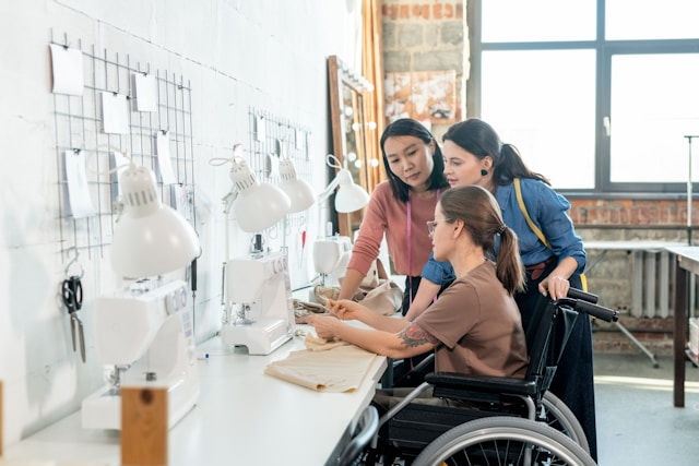 Three women in a sewing room, two standing and one in a wheelchair, discussing by sewing machines. Bright, industrial setting.