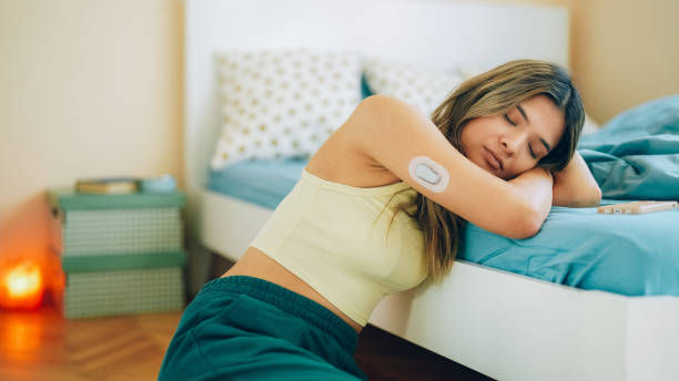 Woman resting on bed edge, wearing a tank top and a glucose monitor on her arm. Bedroom with teal bedding, candlelit ambiance.