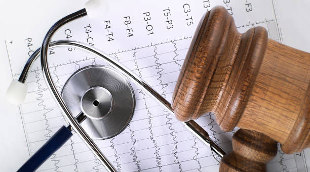 A stethoscope and wooden gavel rest on a sheet with an EKG pattern, blending medical and legal themes. Neutral colors dominate.