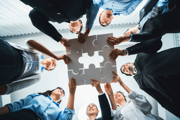 A group of people in business attire collaboratively hold up interconnected puzzle pieces in a bright room, smiling and looking upward.