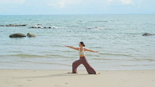 A woman practices yoga in warrior pose on a sandy beach by the ocean. She's wearing a white top and brown pants, exuding calmness and focus.