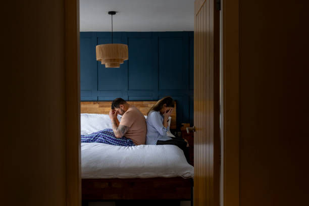 A couple sits back-to-back on a bed, both appearing upset. The room features blue walls, a wood headboard, and a hanging light fixture.