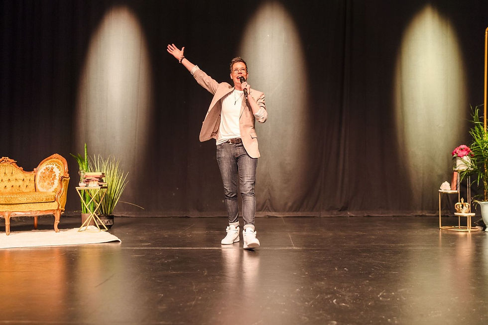 Person excitedly speaking into a microphone on stage, with one arm raised. Background features a vintage sofa, plants, and spotlight.