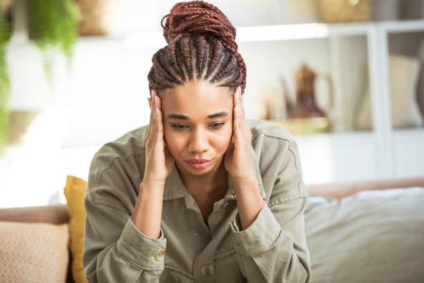 A young woman with braided hair sits indoors, looking stressed or overwhelmed, with her hands pressed to her temples.