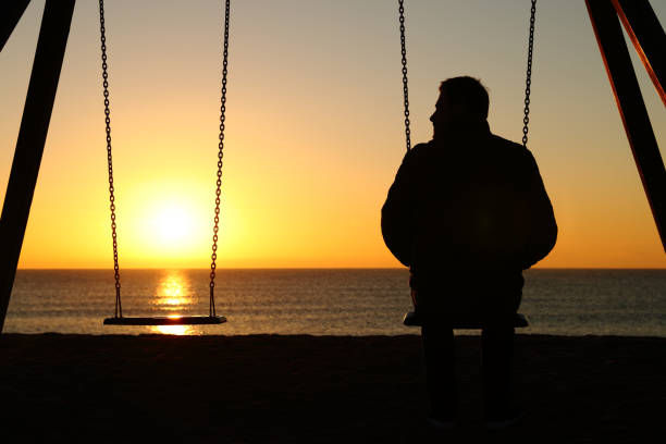 Silhouette of a person sitting on a swing, facing a serene ocean sunset. The sky is orange, creating a peaceful, contemplative mood.