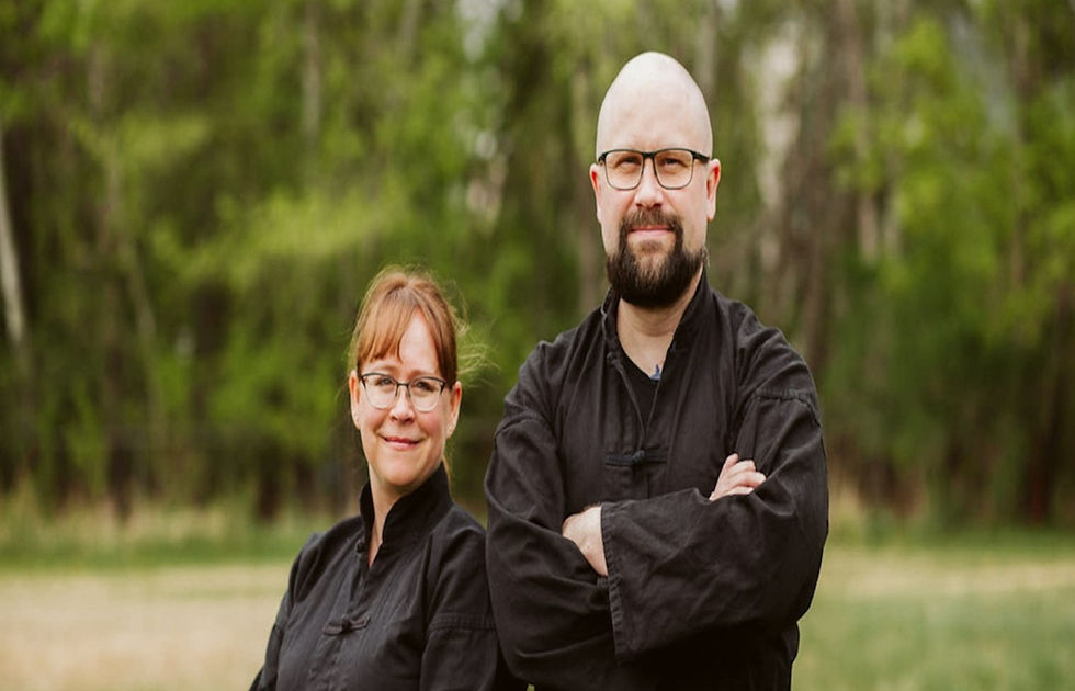 Two people in black clothing stand confidently in a lush green outdoor setting, both smiling calmly. Background features trees and open space.