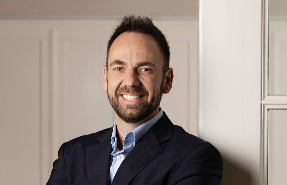 Smiling man in a suit leans against a light-colored wall. He exudes a professional and confident demeanor in a bright, neutral setting.