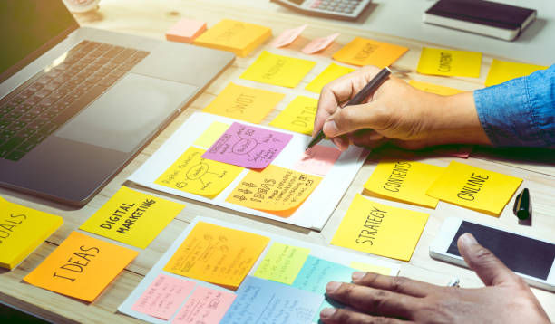 Hands writing on colorful sticky notes on a desk beside a laptop. Notes include "Digital Marketing" and "Strategy." Bright, focused mood.