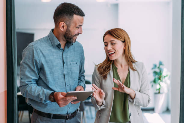 Man and woman in an office setting, smiling and talking. The man holds a tablet. Bright interior with plants in the background.