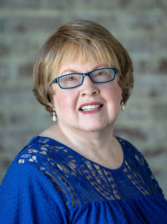 Smiling woman with blue glasses and earrings in a royal blue lace top, posed against a blurred brick wall.