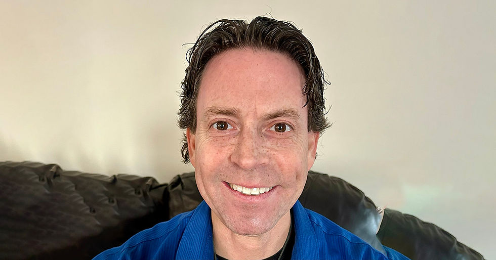 Smiling person with wavy hair in a blue shirt sits on a dark leather couch. Neutral beige background. Appears happy and relaxed.