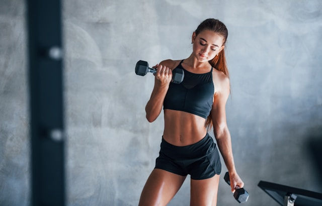 Woman in black athletic wear lifts dumbbells in a gym, focused expression. Gray concrete wall background. Strength and determination theme.