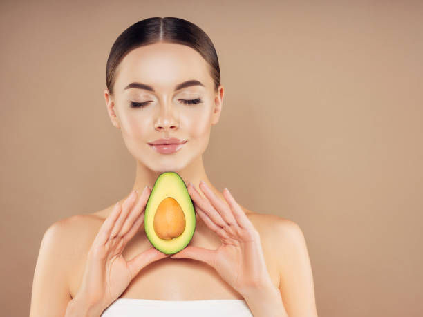 Woman in a white top holding half an avocado near her face with closed eyes, in a serene mood. Beige background enhances calmness.