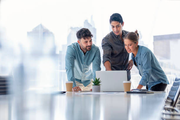 Four people in casual attire discussing in an office near a desk with a computer. One holds papers, another a mug. Bright, modern setting.