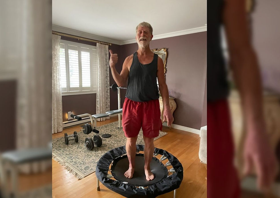 Older man gives thumbs up while standing on a mini trampoline in a home gym living room with dumbbells and bench.