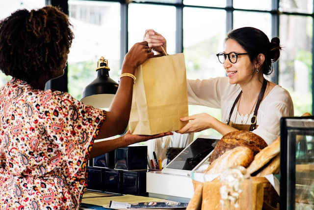 Woman in a bakery smiling, handing a paper bag to a customer. Bread and rolls on the counter. Bright, airy setting with large windows.