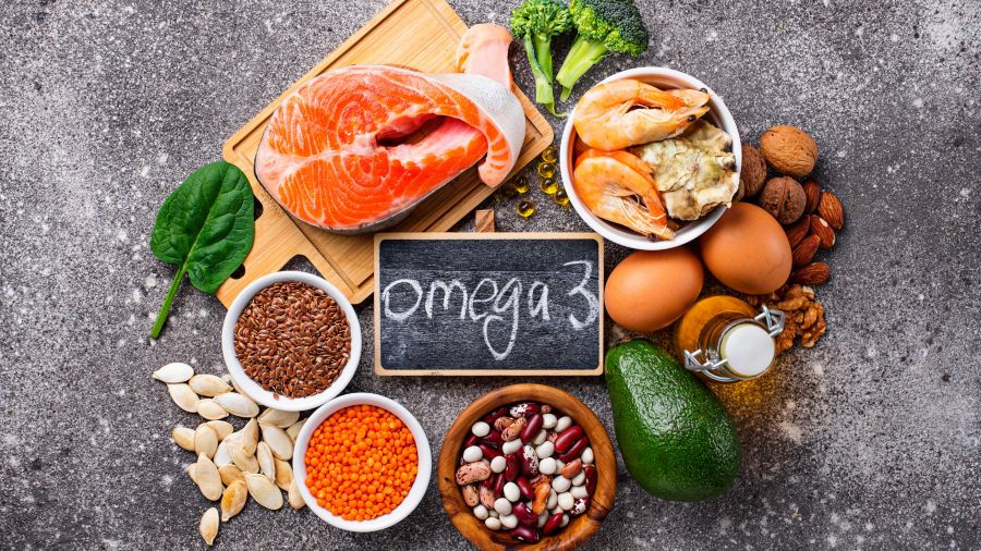The photo displays a variety of omega-3-rich foods, including salmon, shrimp, avocado, nuts, seeds, eggs, and legumes, with a small chalkboard labeled "omega 3" in the center.