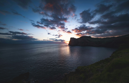 Sunset over a calm sea with dramatic clouds. A silhouette of a cliff on the horizon. Sky colors mix deep blue, pink, and orange. Peaceful mood.