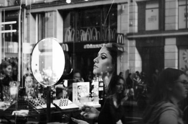 A woman applies makeup in front of a mirror in a store window. Reflections show a busy street with people passing by and a McDonald's sign.