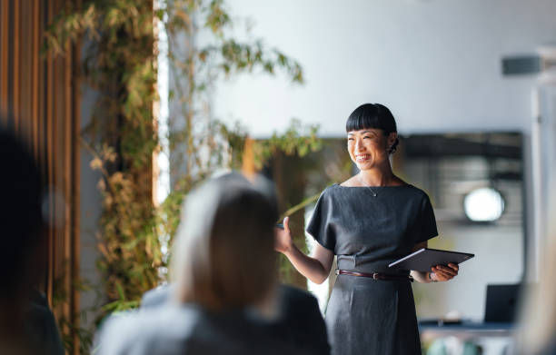 Woman in gray dress smiling while holding a tablet, speaking to an audience. Background includes plants and soft lighting, creating a warm mood.