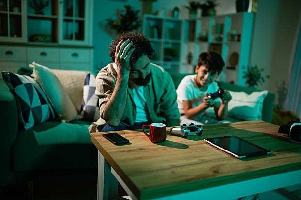 Stressed man on couch with hand on head while boy plays a game behind him in a teal-lit living room, with controller on table