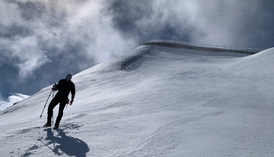 Lone skier climbs a steep snowy mountain slope with poles under dramatic cloudy sky, fenced ridge above.
