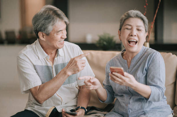 Elderly couple laughing on a sofa, woman holding a smartphone. The man points playfully. Cozy home setting with neutral colors.