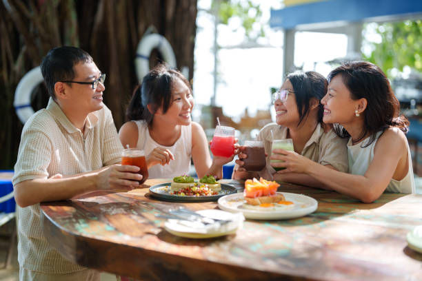 Four people sit at a wooden table outdoors, smiling and holding colorful drinks. Plates of food are on the table. Sunny and cheerful mood.