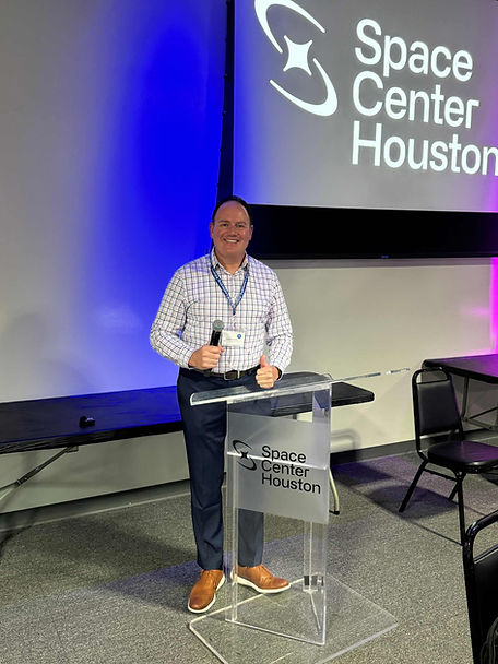 Man smiling at a clear podium with "Space Center Houston" logo, under blue and purple lighting in a conference room.