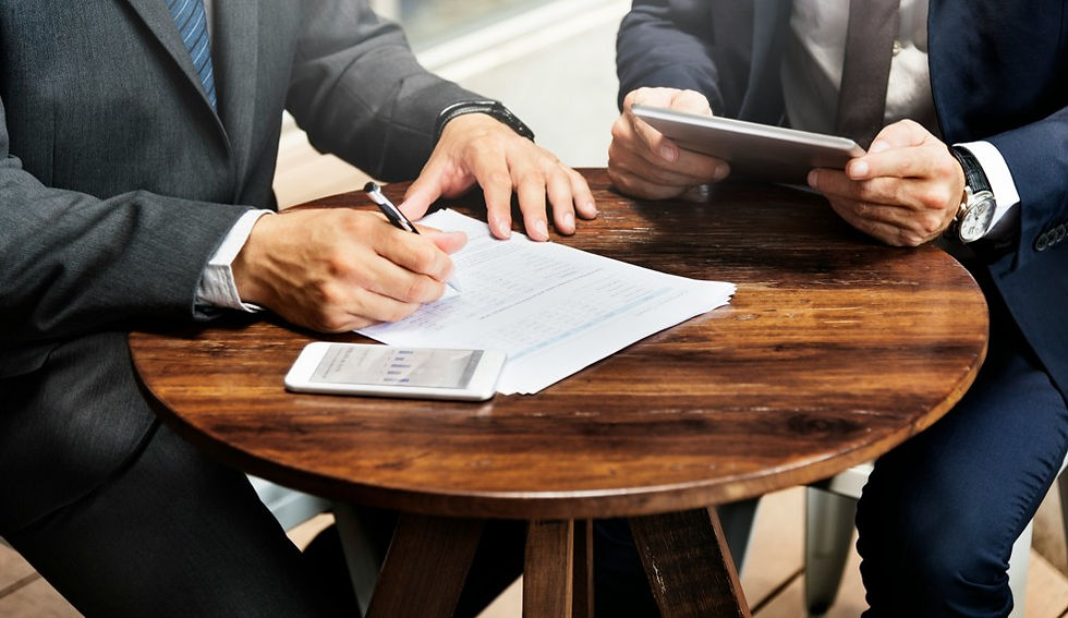 Two suited businessmen review and sign documents at a wooden table, with a tablet and smartphone in a bright office setting.
