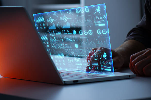 A person interacts with a laptop projecting futuristic blue holographic data charts in a dimly lit room, creating a tech-focused atmosphere.