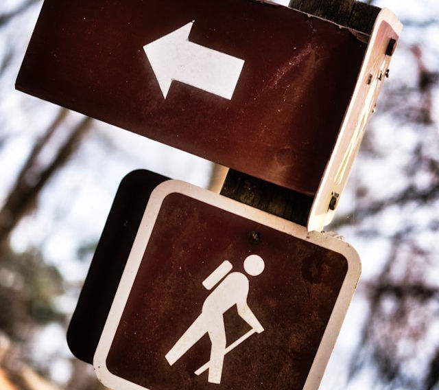 The photo shows two brown trail signs: one with a white arrow pointing left, and another below it with a hiker icon, indicating a nearby hiking trail.