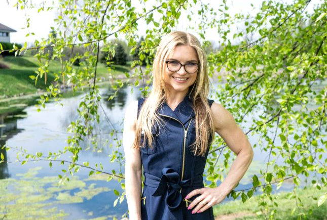Smiling blonde woman in glasses and a navy dress stands by a pond with green trees, in a bright outdoor setting.