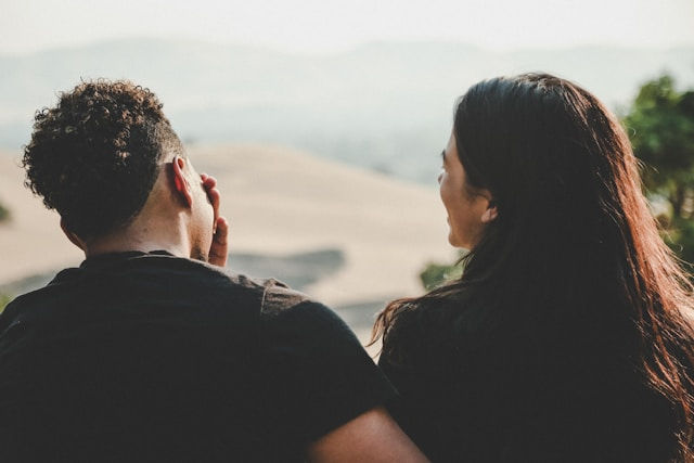 A couple sits outdoors overlooking a blurred scenic landscape, with one person covering their face while the other turns toward them, suggesting a quiet and emotional conversation.