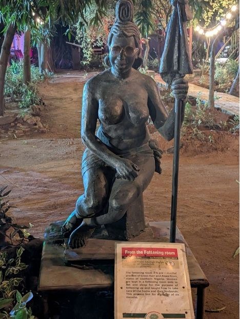 A bronze statue of a woman sitting on a platform, holding a staff, surrounded by greenery. A sign reads "From the Fattening room."