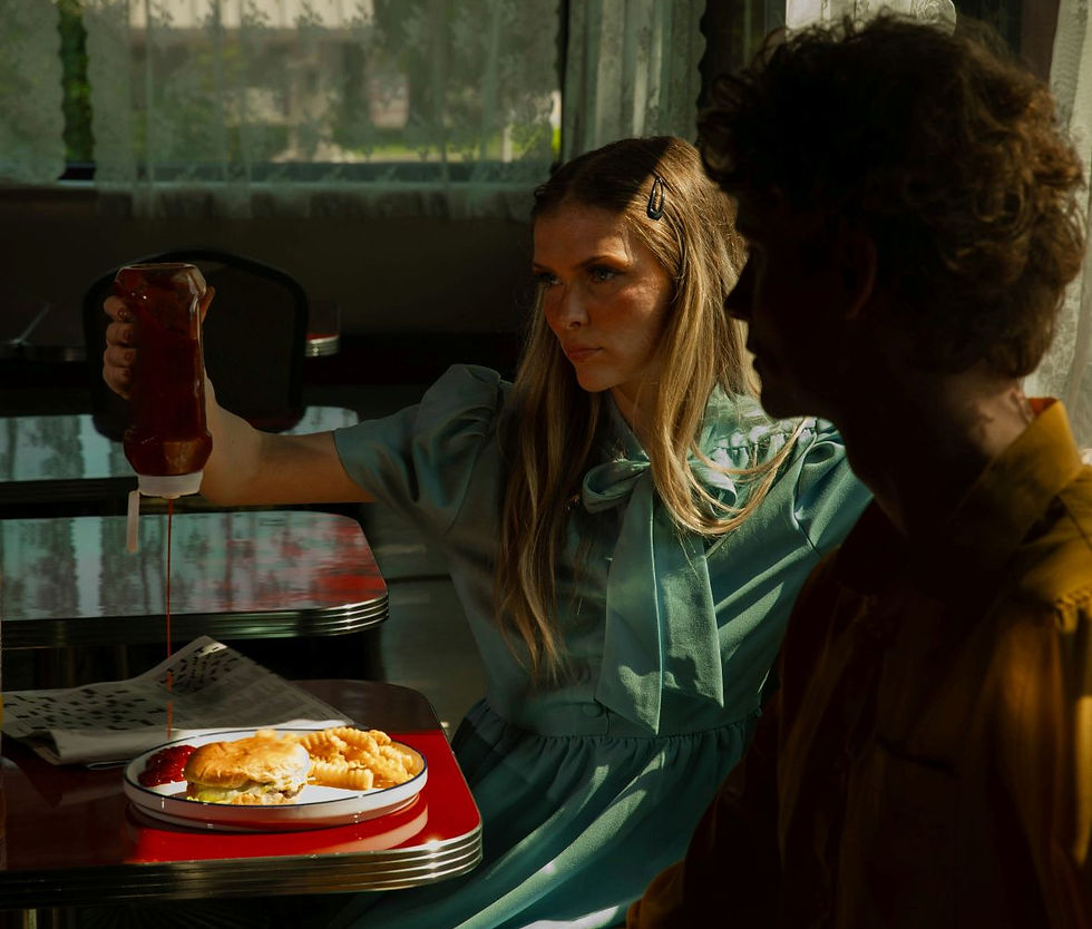 Woman in teal dress pouring ketchup on a plate with a burger and fries, seated in a vintage diner. Sunlit curtains in the background.