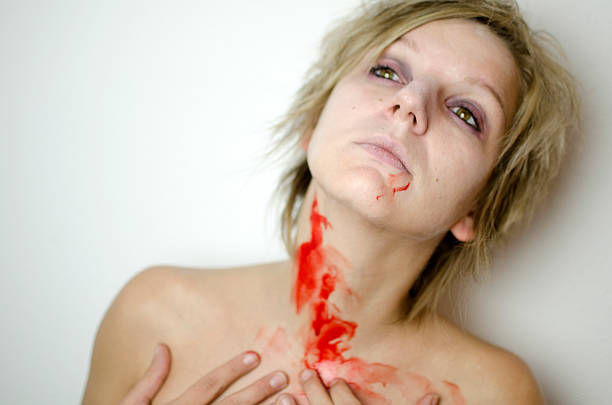 A person with short hair and dramatic eye makeup looks upward, with red paint or liquid on their neck and hand, against a plain white background.