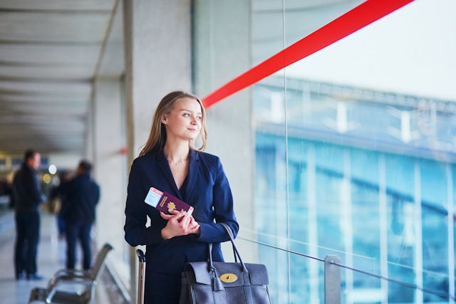 A woman in formal attire standing at an airport terminal, holding a passport and boarding pass, looking thoughtfully out the window.