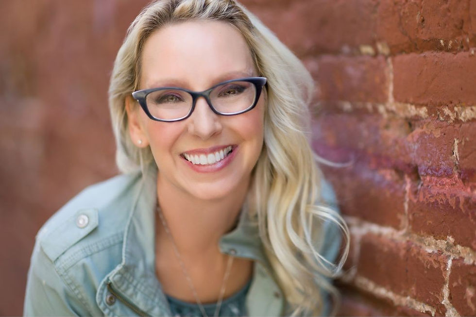 Woman with glasses and blonde hair smiles brightly, leaning against a red brick wall. She wears a light blue jacket, exuding a cheerful vibe.