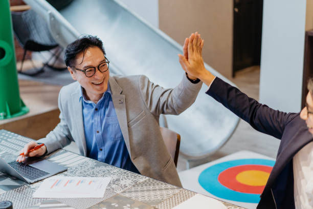 Two people high-fiving in an office with a slide and target pattern mat in the background. The mood is positive and energetic.